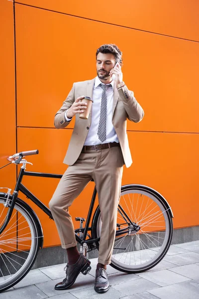 В полный рост бизнесмен разговаривает по смартфону и держит бумажную чашку рядом с велосипедом и зданием с оранжевыми стенами — стоковое фото