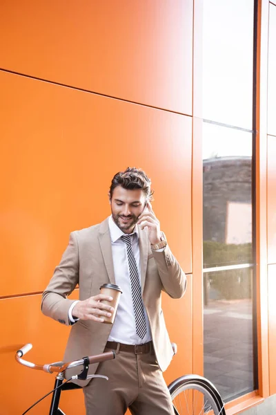 Happy businessman talking on smartphone and holding paper cup near bicycle and building with orange walls — Stock Photo