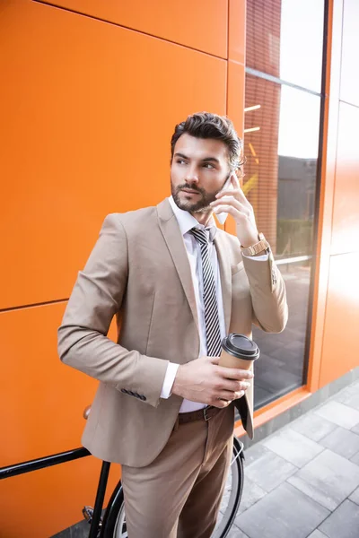 Uomo d'affari fiducioso parlando su smartphone e tenendo tazza di carta vicino alla bicicletta e edificio con pareti arancioni — Foto stock