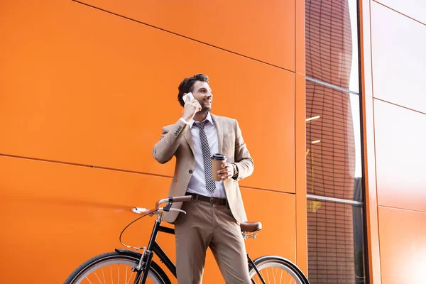 Веселый бизнесмен разговаривает на смартфоне и держит бумажную чашку возле велосипеда и здания с оранжевыми стенами — стоковое фото