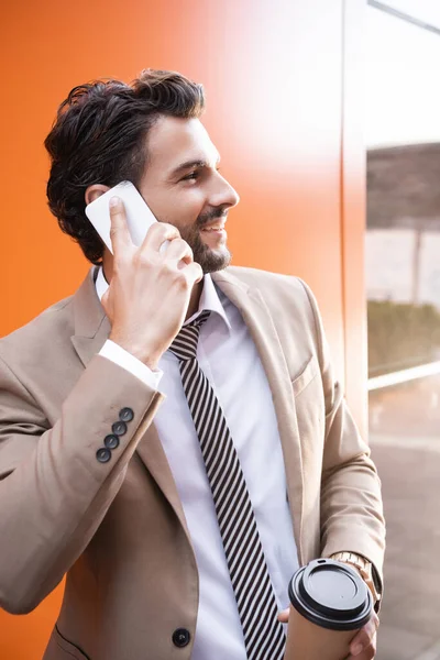 Веселый бизнесмен разговаривает на смартфоне и держит бумажную чашку возле здания с оранжевыми стенами — стоковое фото