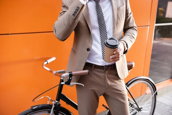 Vue partielle d'un homme d'affaires tenant une tasse en papier près d'un vélo et d'un bâtiment aux murs orange — Photo de stock