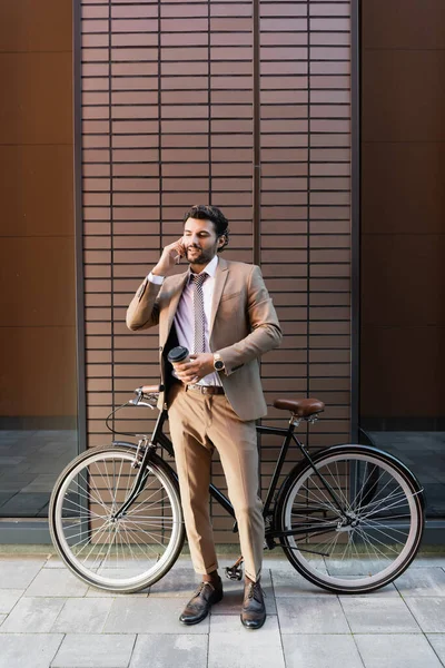 Повна довжина бородатого бізнесмена, який говорить на смартфоні і тримає паперовий стакан біля велосипеда і сучасної будівлі — стокове фото