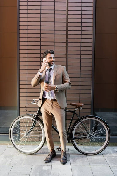 На всю длину бородатый бизнесмен разговаривает на смартфоне и держит бумажную чашку возле велосипеда и здания — стоковое фото