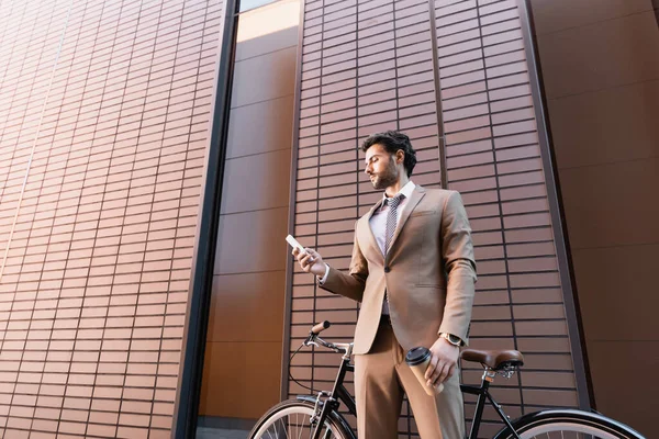 Homme d'affaires barbu regardant smartphone et tenant tasse de papier près de vélo tout en se tenant près du bâtiment — Photo de stock