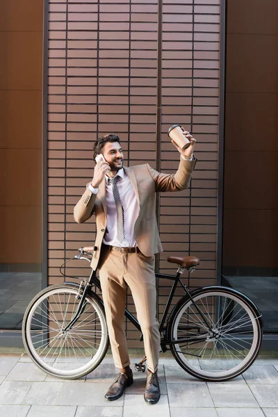 Piena lunghezza di felice uomo d'affari parlando su smartphone e tenendo tazza di carta vicino a bicicletta e costruzione — Foto stock