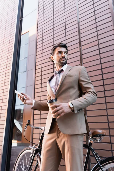 Vue à faible angle de l'homme d'affaires barbu tenant smartphone et tasse en papier près de la bicyclette tout en se tenant près du bâtiment — Photo de stock