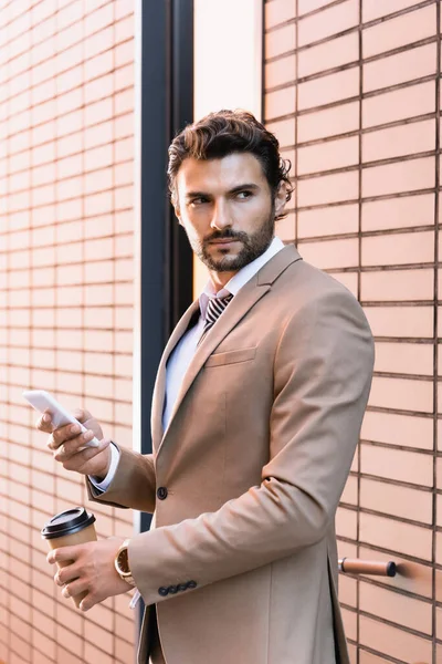 Бородатый бизнесмен, держащий в руках смартфон и бумажную чашку, стоя рядом со зданием — стоковое фото
