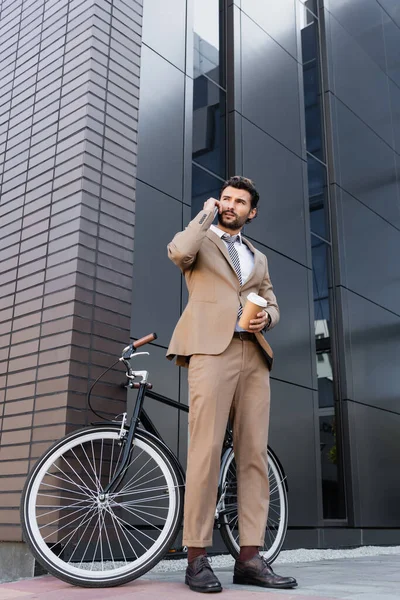 Low angle view of businessman talking on smartphone and holding paper cup near bicycle and building — Stock Photo
