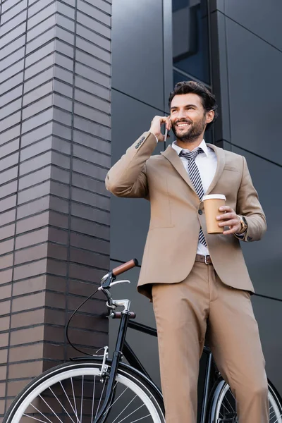 Tiefansicht eines fröhlichen Geschäftsmannes, der mit Smartphone spricht und Pappbecher in der Nähe von Fahrrad und Gebäude hält — Stockfoto