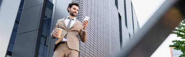 Vista de ángulo bajo del hombre de negocios alegre mirando el teléfono inteligente y sosteniendo la taza de papel cerca del edificio, pancarta - foto de stock