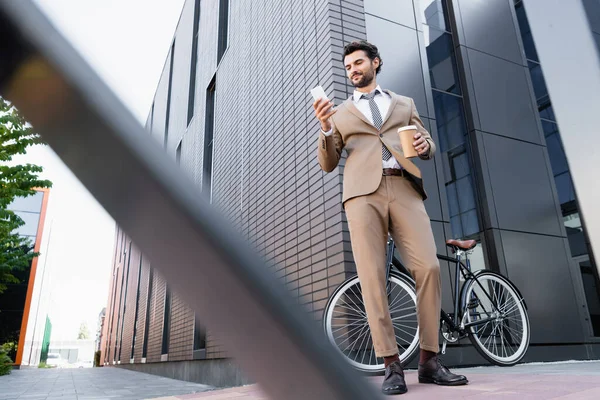 Низкий угол зрения веселого бизнесмена, смотрящего на смартфон и держащего бумажную чашку возле велосипеда и размытого переднего плана — стоковое фото
