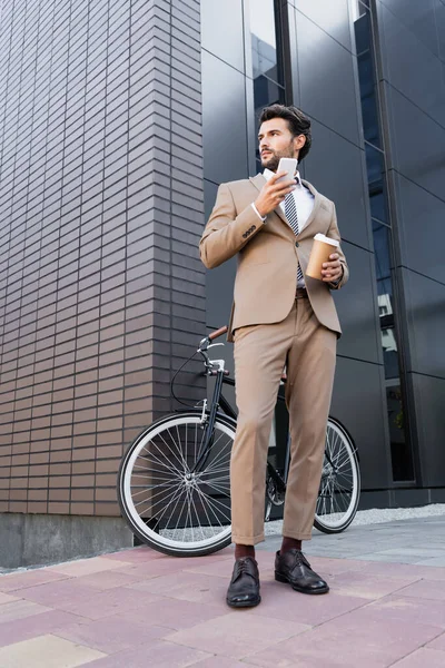 Низкий угол обзора бизнесмена, держащего смартфон и кофе, чтобы идти рядом с велосипедом и зданием — стоковое фото