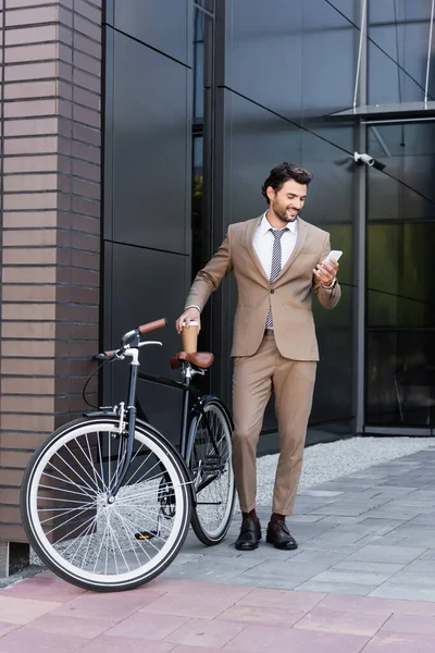 Pleine longueur d'homme d'affaires joyeux tenant smartphone et café pour aller près de vélo tout en se tenant près du bâtiment — Photo de stock