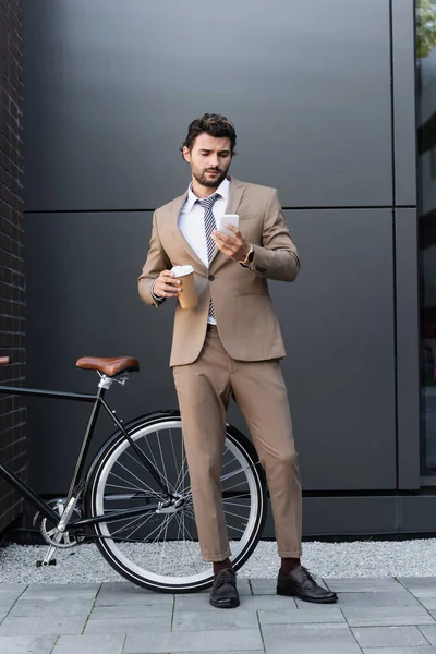 Toute la longueur de barbu homme d'affaires tenant café pour aller et regardant smartphone près de vélo — Photo de stock