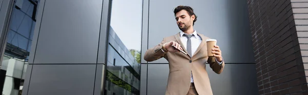 Bearded businessman holding coffee to go and smartphone while looking at watch near building, banner — Stock Photo