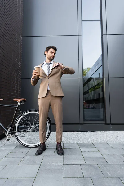 Longitud completa de hombre de negocios barbudo en traje sosteniendo café para ir y teléfono inteligente mientras mira el reloj cerca de la bicicleta - foto de stock