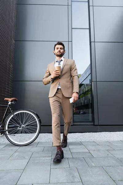 Pleine longueur de barbu homme d'affaires en costume tenant café pour aller et smartphone tout en marchant près de vélo — Photo de stock