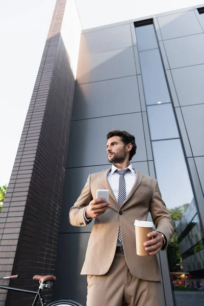Vista de ángulo bajo del hombre de negocios barbudo en traje sosteniendo café para llevar y teléfono inteligente cerca de edificio moderno - foto de stock