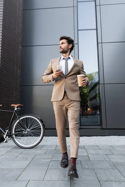 Full length of businessman in suit holding coffee to go and smartphone while walking near bicycle — Stock Photo