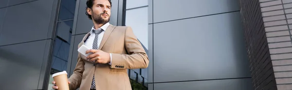 Uomo d'affari barbuto in abbigliamento formale che tiene il caffè per andare e smartphone vicino all'edificio moderno, banner — Foto stock