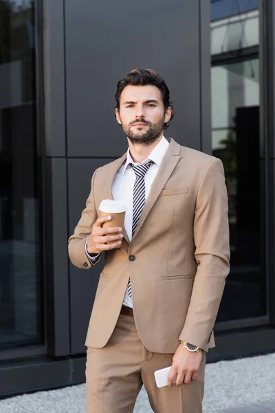 Hombre de negocios barbudo en ropa formal sosteniendo café para llevar y teléfono inteligente cerca de edificio moderno - foto de stock