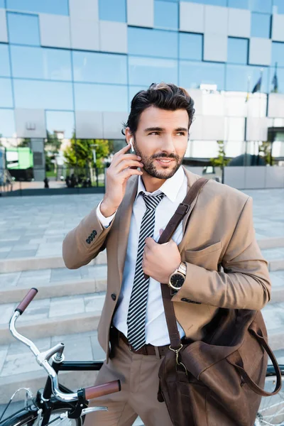 Bearded businessman adjusting wireless earphones while standing near bicycle — Stock Photo