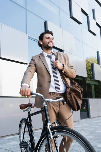 Cheerful businessman in wireless earphones while standing with bag near bike and building — Stock Photo