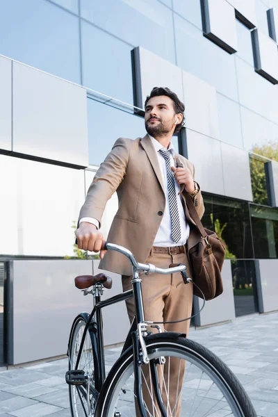 Businessman in wireless earphones while standing with bag near bike and building — Stock Photo