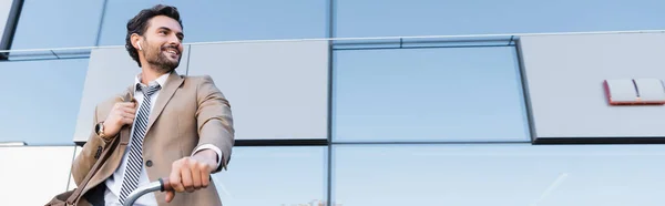 Low angle view of cheerful businessman in wireless earphones while standing with bag near bike and building, banner — Stock Photo