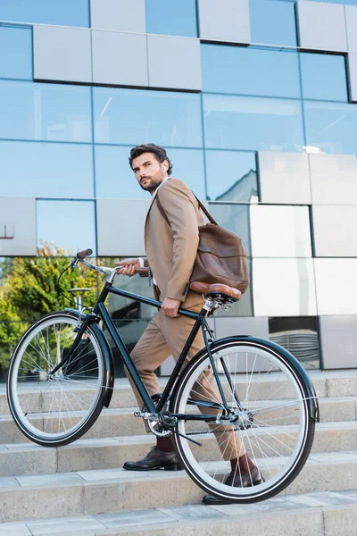 Homem de negócios sério em fones de ouvido sem fio andando em escadas com bicicleta — Fotografia de Stock