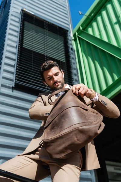 Vista de ángulo bajo del hombre de negocios barbudo en auriculares inalámbricos que sostienen el bolso de cuero - foto de stock