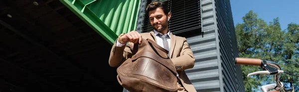 Vista de ángulo bajo de empresario feliz en auriculares inalámbricos que sostienen el bolso de cuero, bandera - foto de stock