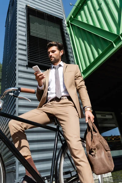 Vue à faible angle de l'homme d'affaires barbu dans les écouteurs sans fil debout près du vélo et tenant smartphone — Photo de stock