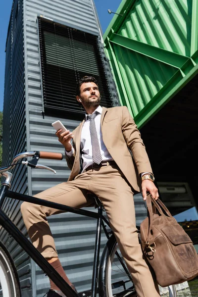 Vue à faible angle de l'homme d'affaires dans les écouteurs sans fil debout près de vélo et tenant smartphone — Photo de stock