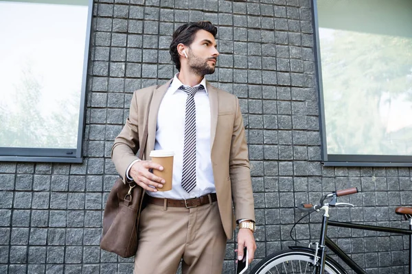 Homme d'affaires dans les écouteurs sans fil et costume tenant tasse en papier et smartphone près du bâtiment — Photo de stock