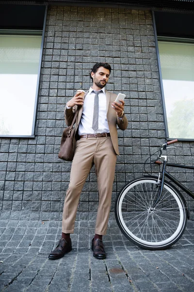 В полный рост бизнесмен в беспроводных наушниках и костюме держит бумажную чашку и смартфон возле здания — стоковое фото
