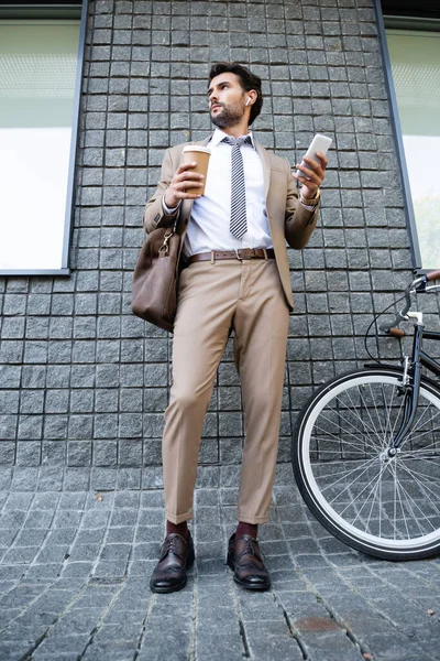 Longitud completa de hombre de negocios en auriculares inalámbricos sosteniendo taza de papel y teléfono inteligente cerca del edificio - foto de stock