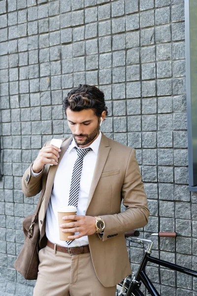 Businessman in wireless earphones and formal wear holding coffee to go and smartphone near building — Stock Photo