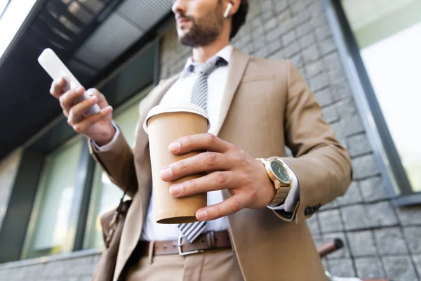 Cropped view of businessman in wireless earphones and suit holding coffee to go and smartphone near building — Stock Photo