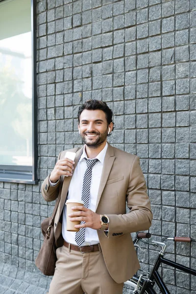Щасливий бізнесмен в бездротових навушниках і костюмі, що тримає каву, щоб піти і смартфон біля будівлі — стокове фото