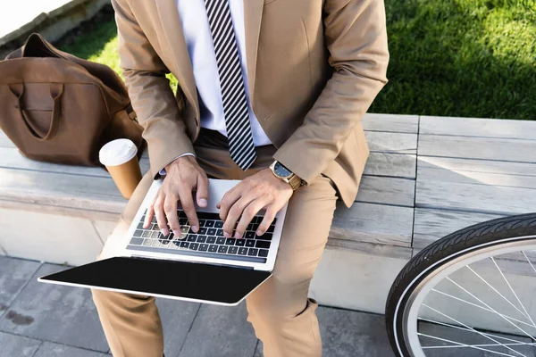 Vista parziale di uomo d'affari utilizzando il computer portatile vicino tazza di carta e seduto sulla panchina — Foto stock