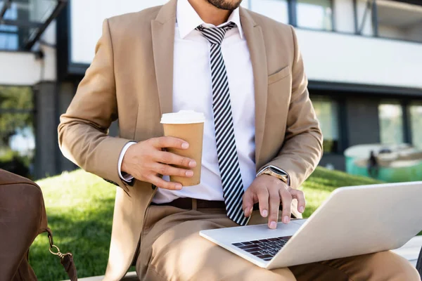 Vista recortada del hombre de negocios utilizando el ordenador portátil y la celebración de la taza de papel - foto de stock
