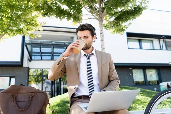 Homem de negócios em fones de ouvido sem fio e terno beber café para ir e usar laptop enquanto sentado no banco — Fotografia de Stock