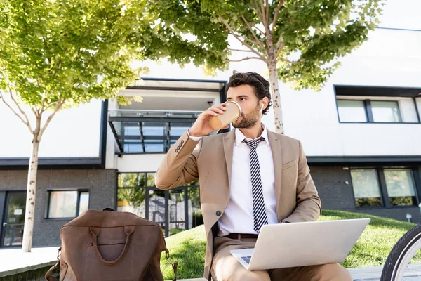 Bearded businessman in wireless earphones and suit drinking coffee to go and using laptop while sitting on bench — Stock Photo