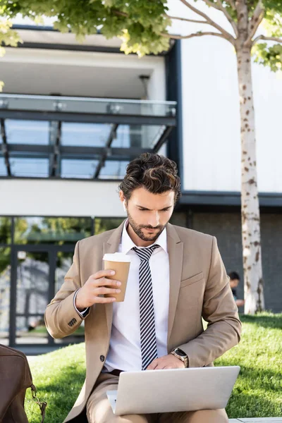 Бізнесмен в бездротових навушниках і костюм тримає каву, щоб піти і використовувати ноутбук, сидячи на лавці — стокове фото