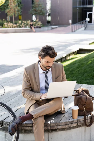 Hombre de negocios en traje usando el ordenador portátil mientras está sentado en el banco con la taza de papel y la bolsa - foto de stock