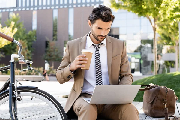 Homem de negócios barbudo em terno usando laptop e segurando copo de papel enquanto sentado no banco perto da bicicleta — Stock Photo