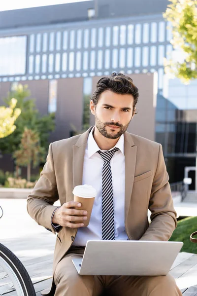 Hombre de negocios en traje sosteniendo taza de papel mientras está sentado en el banco con el ordenador portátil - foto de stock