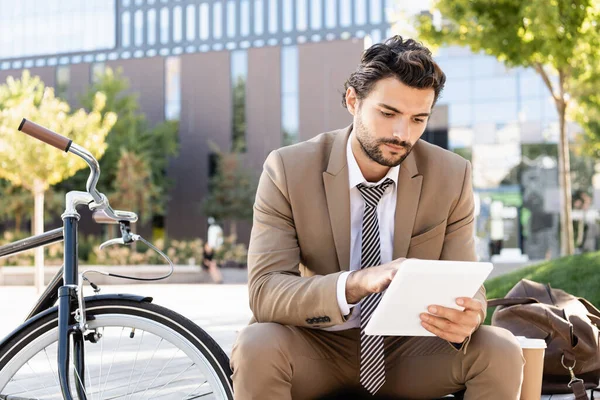 Uomo d'affari in giacca e cravatta con tablet digitale seduto sulla panchina vicino alla bici — Foto stock